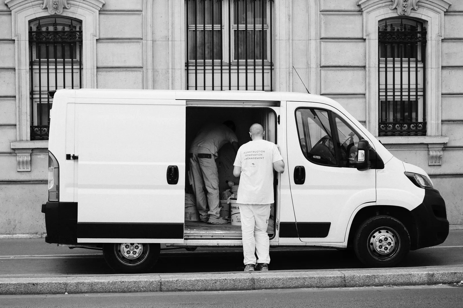 A man loading a white cargo van parked at the curb in front of a stone building